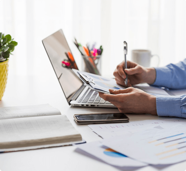Hand holding a pen over a clipboard beside a laptop, smartphone and printed financial charts